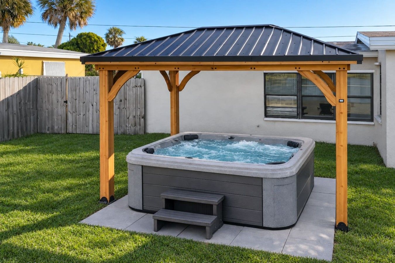 Ein Whirlpool unter einer Holzpergola in einem Garten mit einer weißen Wand und einem Holzzaun im Hintergrund.