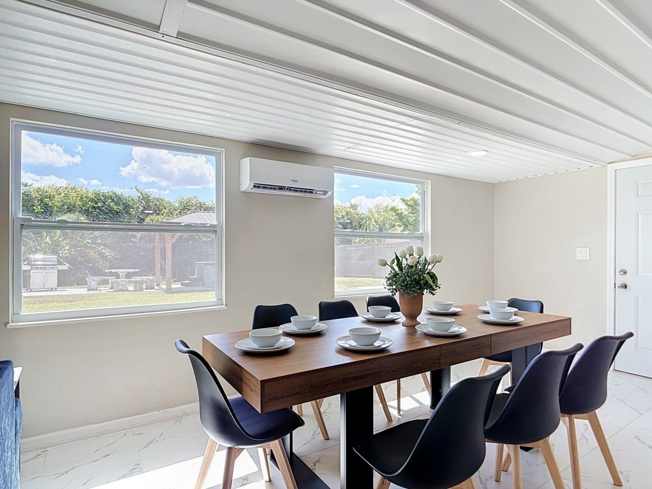 Un coin repas avec une table en bois dressée pour six personnes, entourée de chaises noires, avec une plante en pot sur la table et une vue sur l’extérieur grâce à de grandes fenêtres.