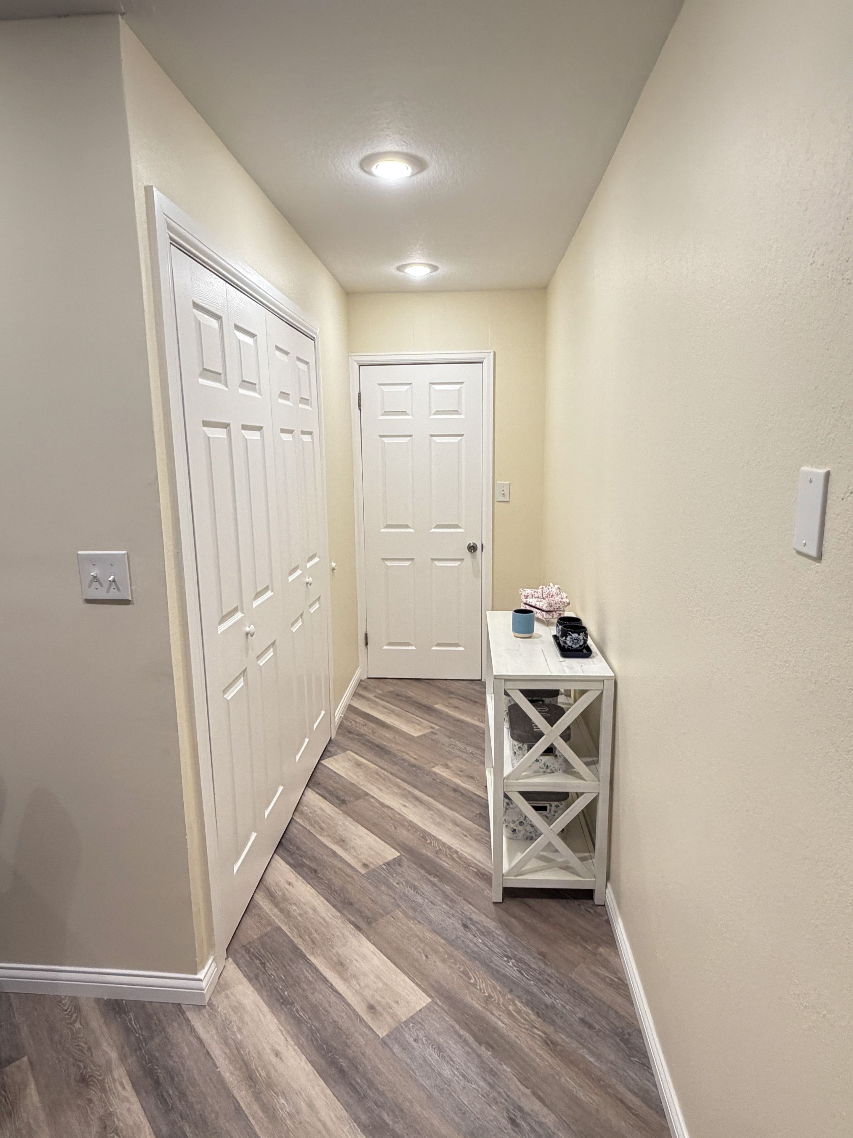 A hallway with a white door, a white table with a vase, and a light switch on the wall.