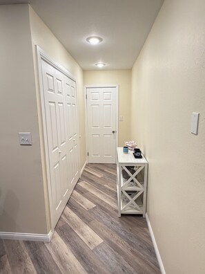 Un pasillo con una puerta blanca, una mesa blanca con un florero y un interruptor de luz en la pared.