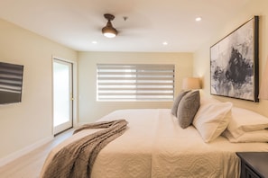 Ein Schlafzimmer mit einem Bett, einem Deckenventilator, einem Fenster mit Jalousien, einem Gemälde an der Wand und einem Fernseher an der Wand.