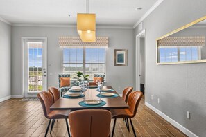 A dining room with a wooden table set for six, surrounded by chairs, with a large window and blinds on the wall. - Vista Cay at Epic Universe : Luxury Lake view 4 bedroom condo (Orlando)