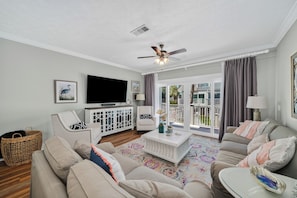 A living room with a flat-screen TV, ceiling fan, and large windows with curtains. - Brand New Listing, Beach, Pool, 2 K Beds (Mexico Beach)