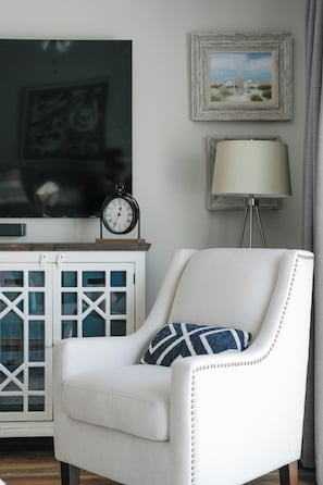 A white armchair with a geometric patterned pillow, a clock, a framed picture, and a floor lamp on a white cabinet. - Brand New Listing, Beach, Pool, 2 K Beds (Mexico Beach)