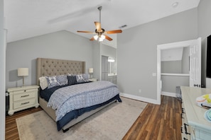Une chambre à coucher avec une tête de lit capitonnée, un lit avec une literie à motifs, une table de chevet avec une lampe, un ventilateur de plafond et un dressing avec un miroir.