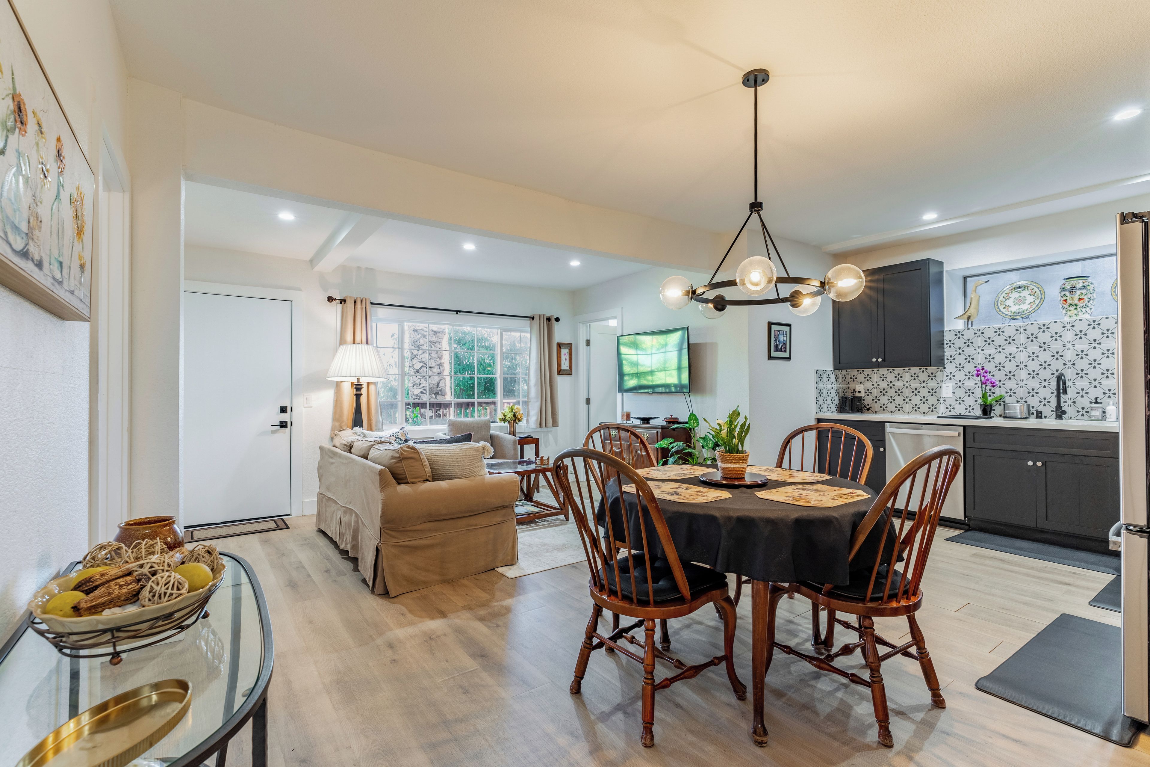 A modern kitchen with dark cabinets, a dining area with a round table and chairs, and a living room with a sofa and a TV.