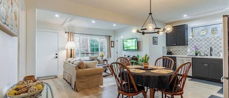 A modern kitchen with dark cabinets, a dining area with a round table and chairs, and a living room with a sofa and a TV.