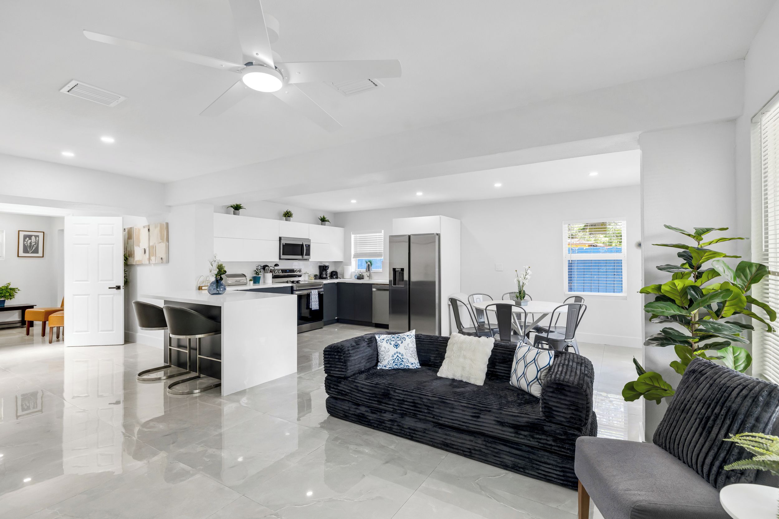 A spacious living area with a black sofa, a grey sofa, a dining table with chairs, and a kitchen with modern appliances.