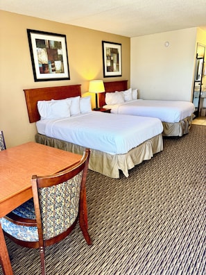 A hotel room with two beds, a wooden table, a patterned chair, and a carpeted floor.