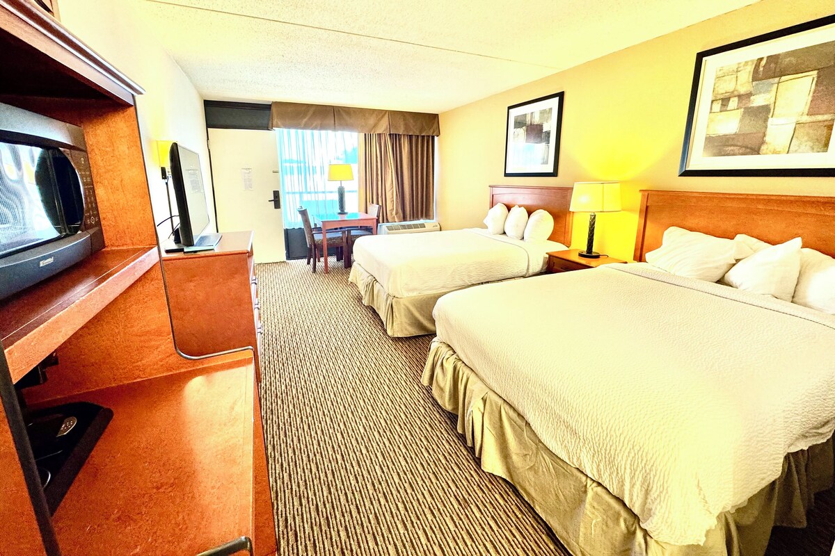 Ett hotellrum med två sängar, ett skrivbord, en stol, en tv och ett fönster med gardiner.