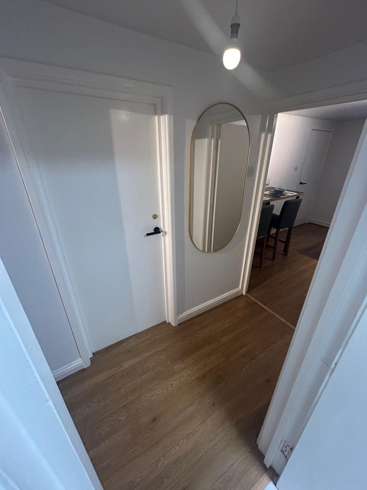 A hallway with wooden flooring, white walls, and a door with a round mirror.