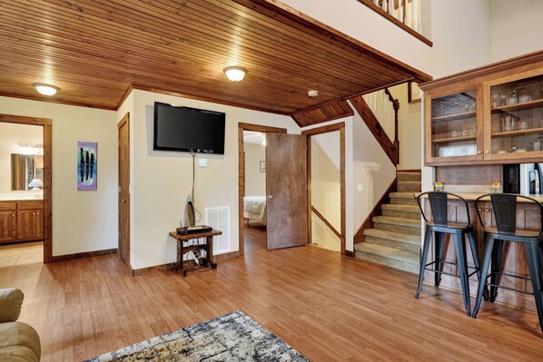 Un vaste espace de vie doté d’un plancher en bois, d’un téléviseur à écran plat fixé au mur, d’un escalier menant à un autre niveau et d’un coin bar avec des tabourets.