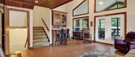 Amplia sala de estar con una escalera de madera, una cocina con taburetes de bar y ventanas grandes.
