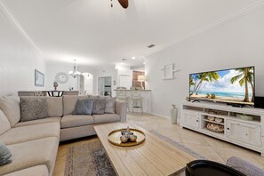 A modern living room with a sectional sofa, a coffee table with a tray, a television on a white cabinet, and a kitchen area in the background. - ⚓Waterview-Resort Style Amenities-Bikes & More⚓ (Bradenton)