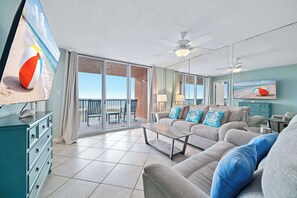 A living room with a sofa, a coffee table, and a television. There is a balcony with a view of the beach. - Buena Vista 201 - Gulf-Front 3BR, Balcony Over the Pool - Beachball Properties (Gulf Shores)