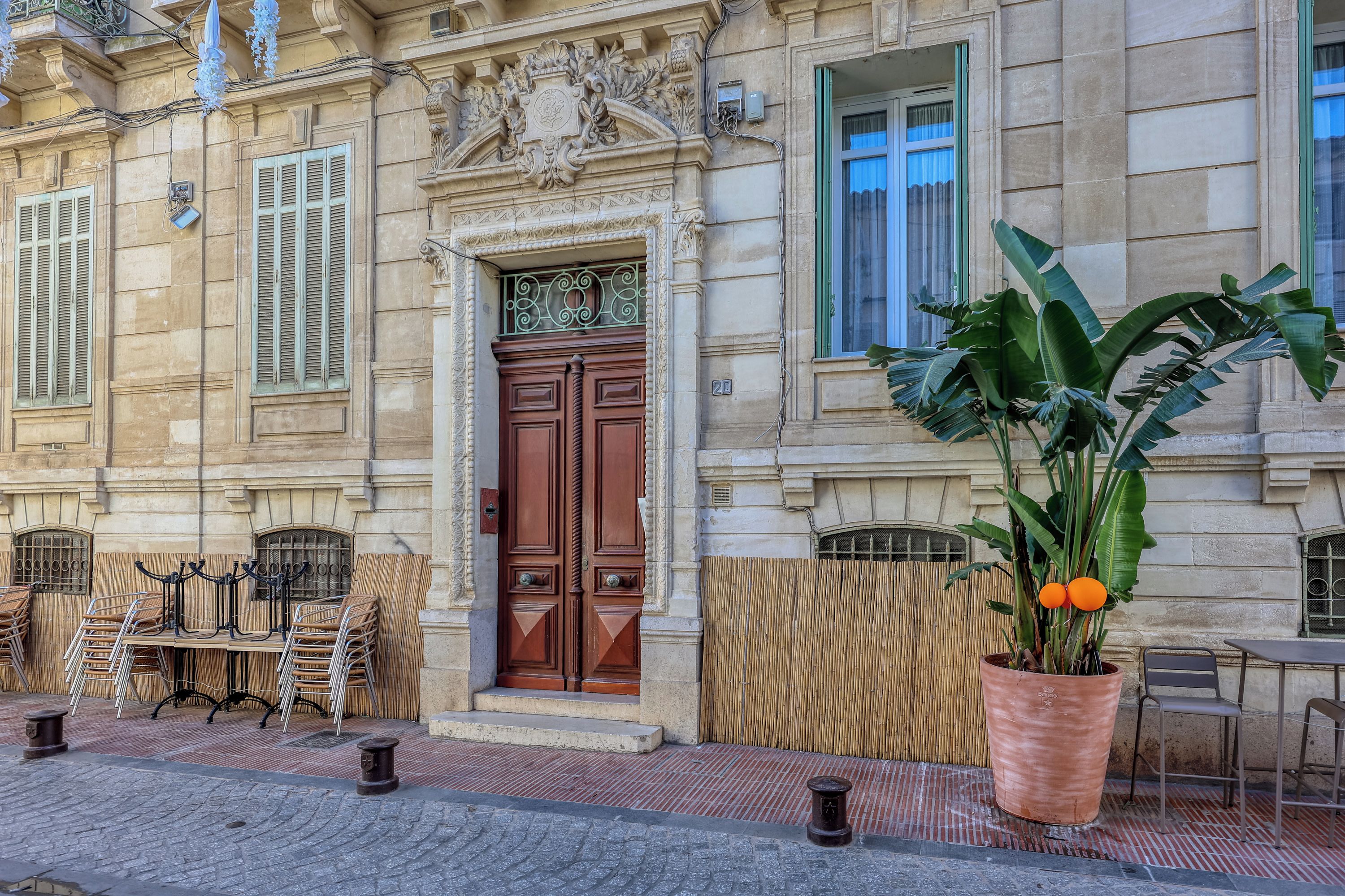 A building facade with a large wooden door, a balcony with a wrought iron railing, and a potted plant with large leaves.