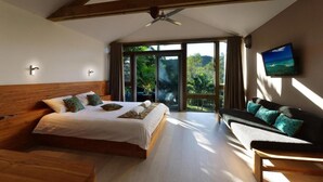 Quarto com cama grande, sofá, televisão e varanda com vista para área verde.