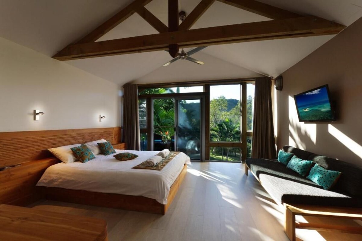 Un dormitorio con una cama grande, un sofá, un televisor y un balcón con vistas a la vegetación.