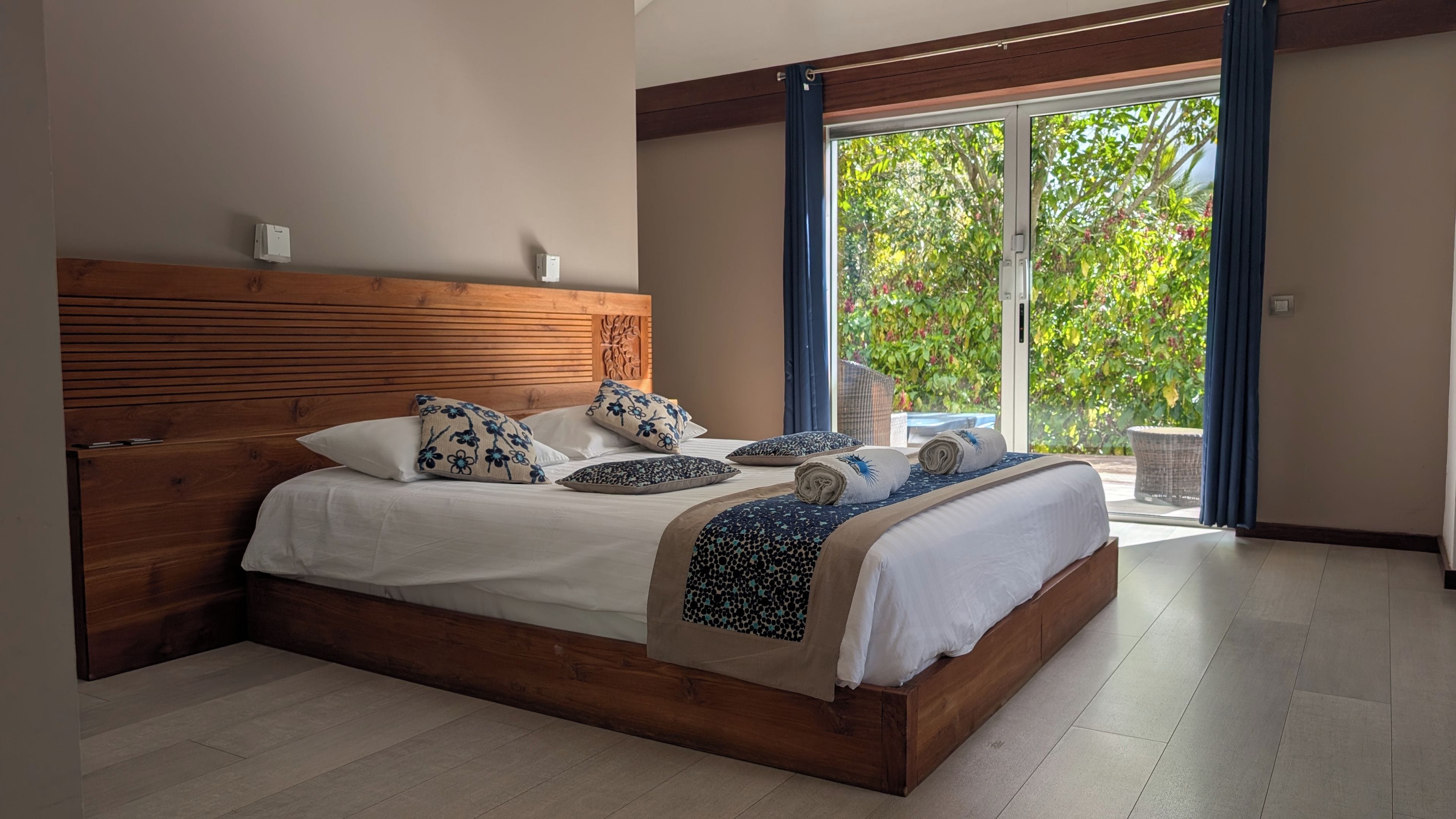 A bedroom with a wooden headboard, a large bed with multiple pillows, and a view of greenery through a sliding glass door.