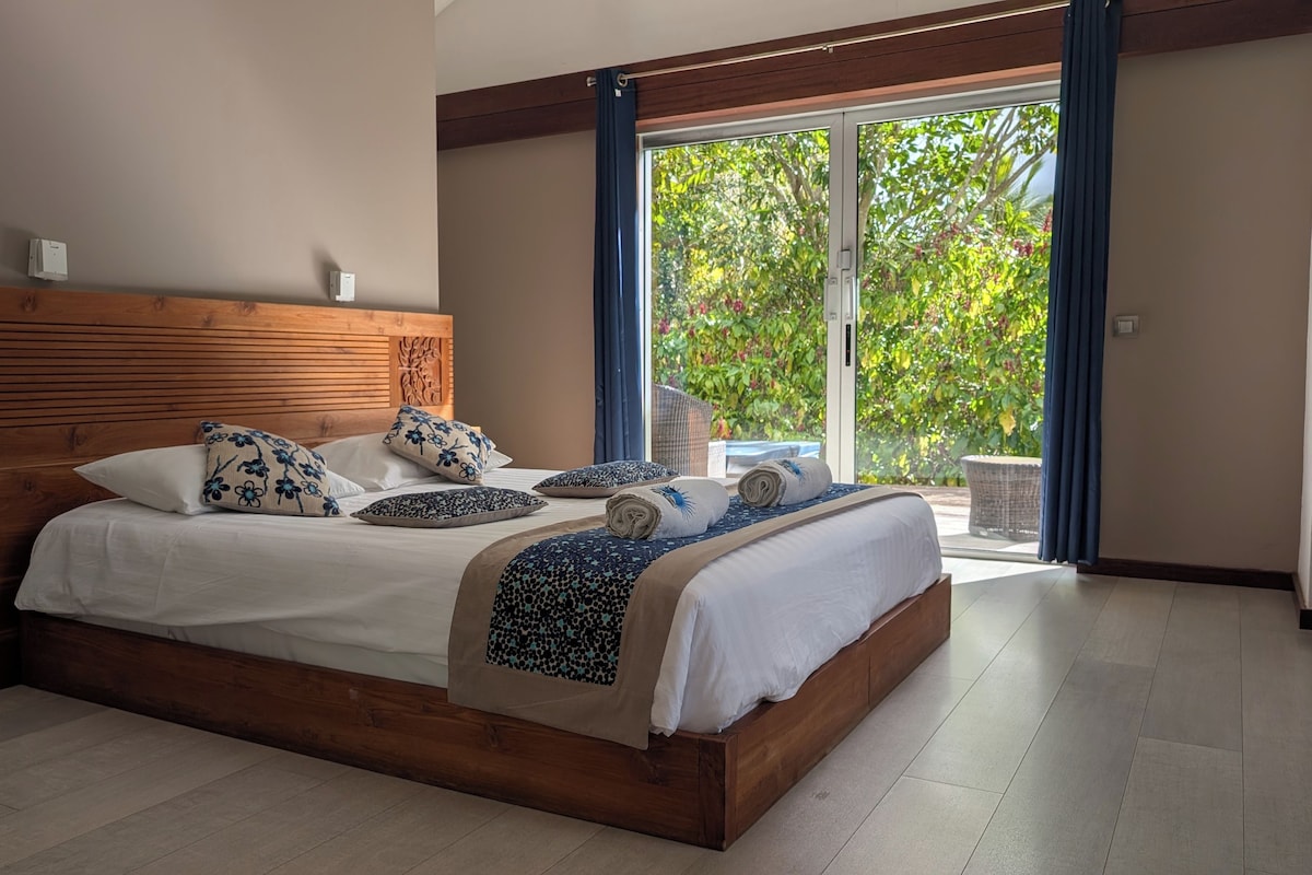A bedroom with a wooden headboard, a large bed with multiple pillows, and a view of greenery through a sliding glass door.