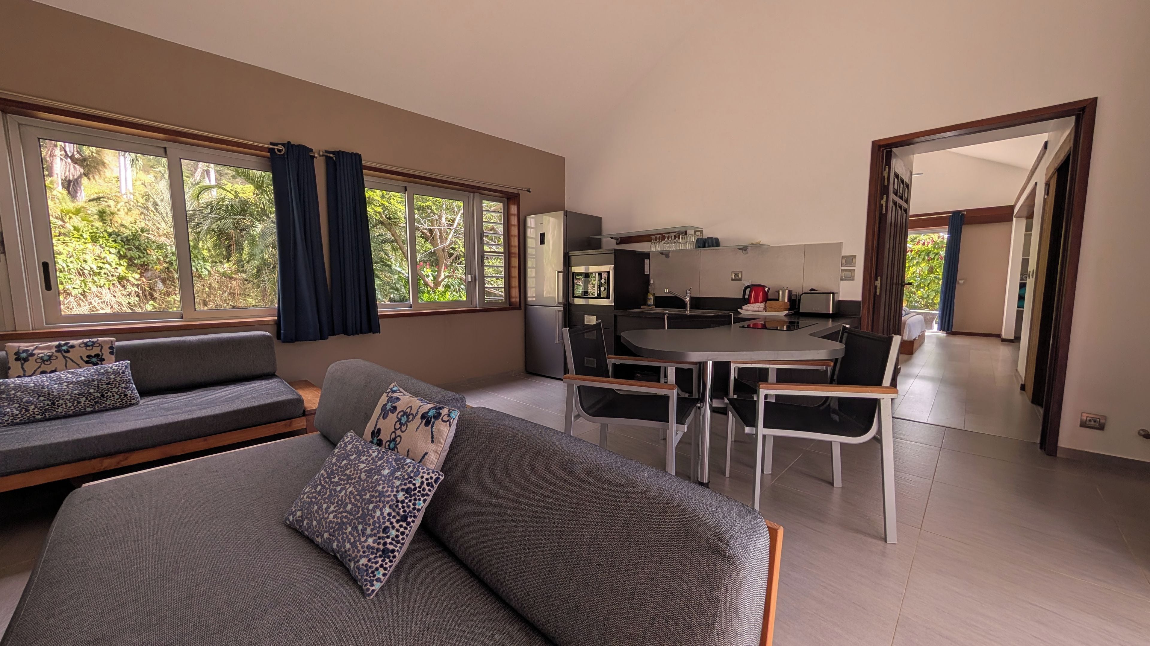 Uma sala de estar moderna com sofá e poltronas, um espaço de jantar com mesa e cadeiras, e uma cozinha ao fundo.