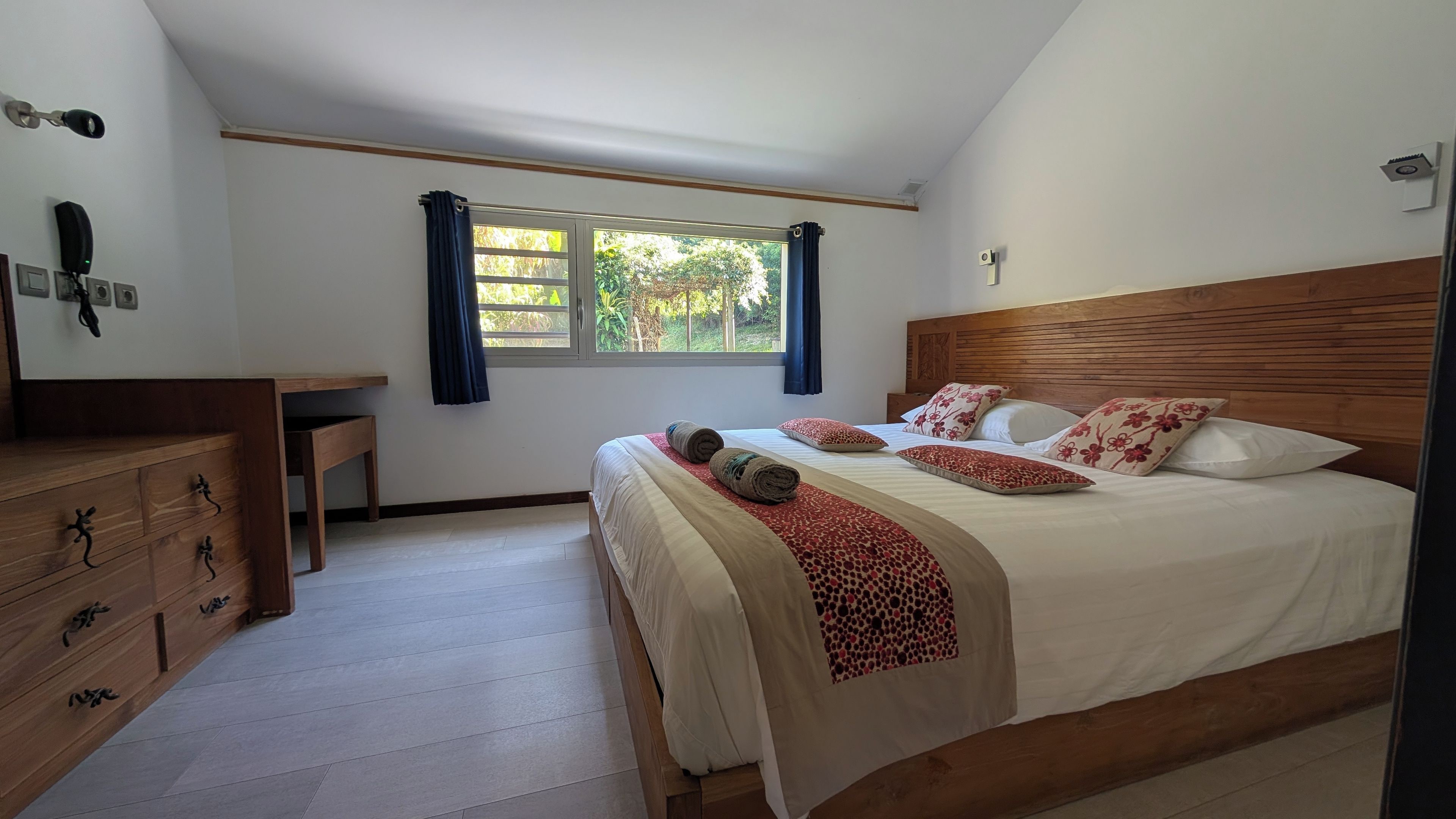 A bedroom with a wooden headboard, a large bed with multiple pillows, a bedside table, and a window with curtains.