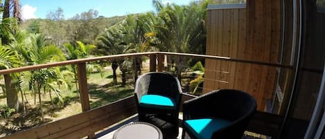 Una terraza de madera con dos sillas de mimbre y una mesa redonda pequeña, con vista a un paisaje tropical con palmeras.