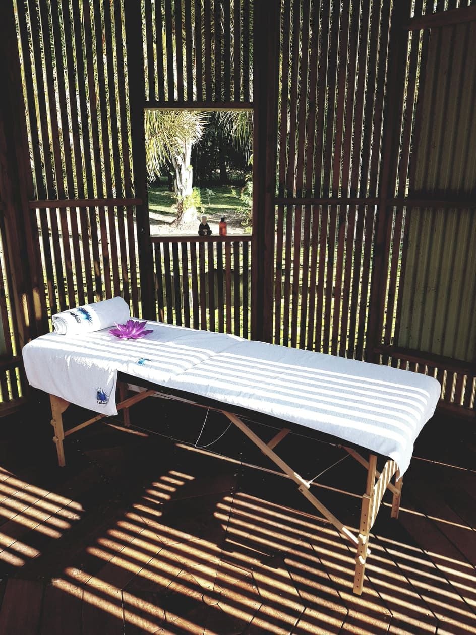 Une table de massage recouverte d’un drap blanc et ornée d’une fleur violette, le tout placé devant un fond en bois composé de lattes verticales.