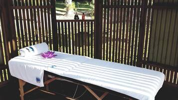 Une table de massage recouverte d’un drap blanc et ornée d’une fleur violette, le tout placé devant un fond en bois composé de lattes verticales.