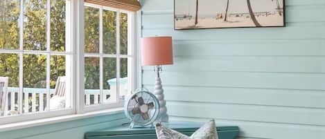 Une pièce avec un mur de couleur turquoise, un fauteuil rose, une petite table ronde sur laquelle se trouve un livre, et un ventilateur.