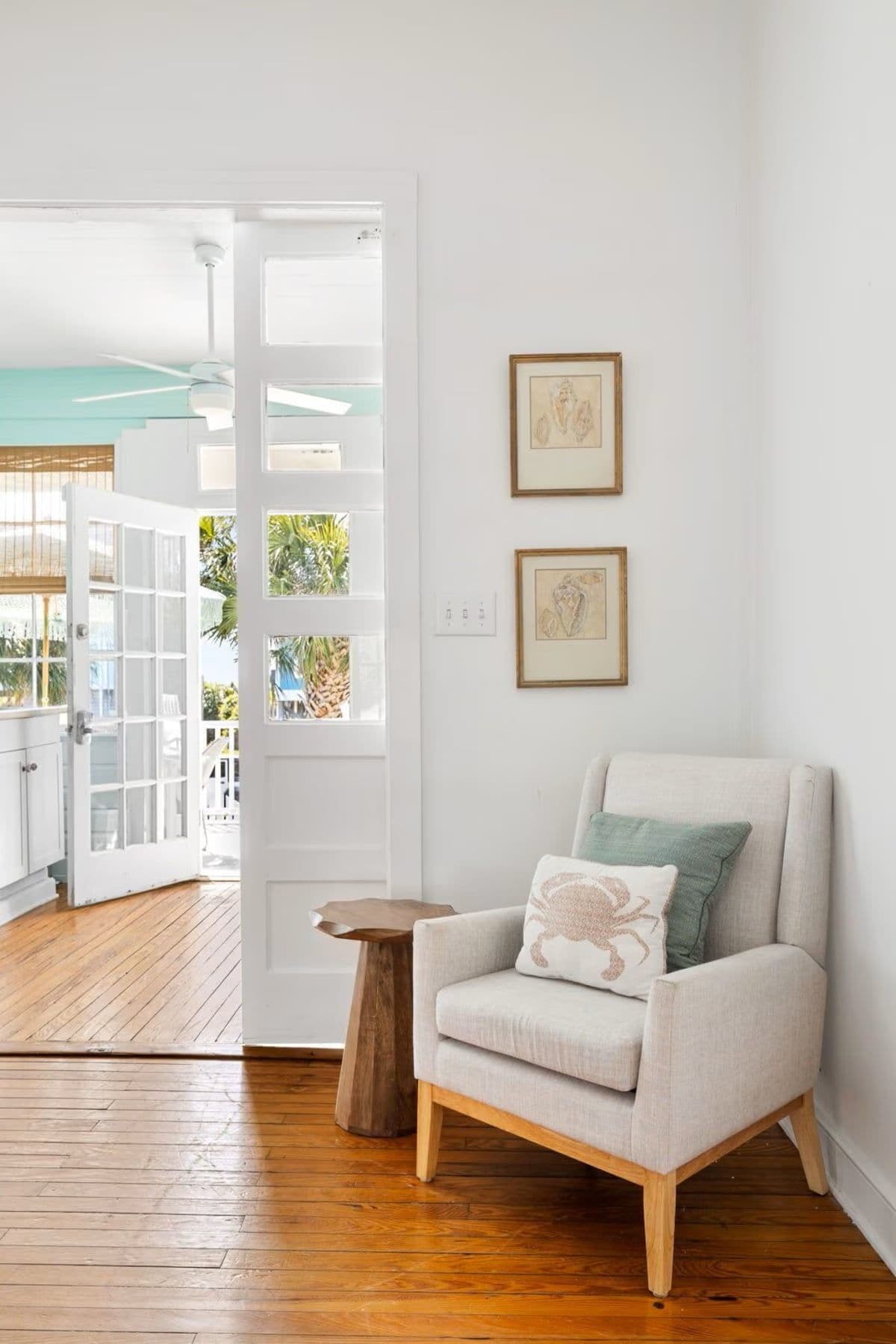 A room with a wooden floor, a white door leading outside, a small wooden side table, and a light-colored armchair with a cushion.