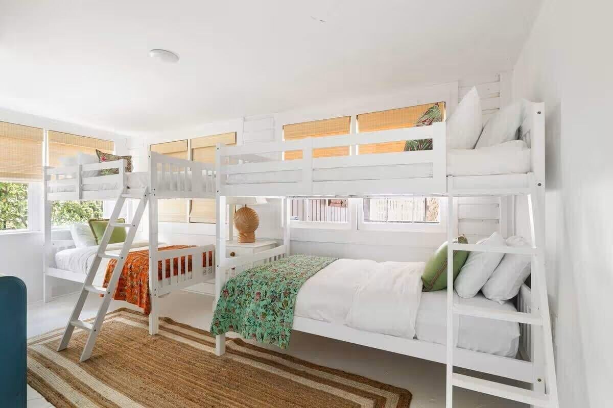 Habitación con literas, una escalera, ventana con persianas y una mesita con un florero.