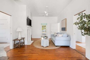 A modern living room with a wooden floor, a light blue sofa, a small round coffee table, a white lamp, and a ceiling fan. - Beachside Bliss | 4BR (plus a bonus bedroom)  Folly Beach Home Steps from Beach Access (Folly Beach)