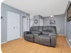 A living room with a grey sofa, a white door, a ceiling fan, and a wall clock. - Graceful Stays: Cozy Highlands 1BR – Unit 6 (Louisville)