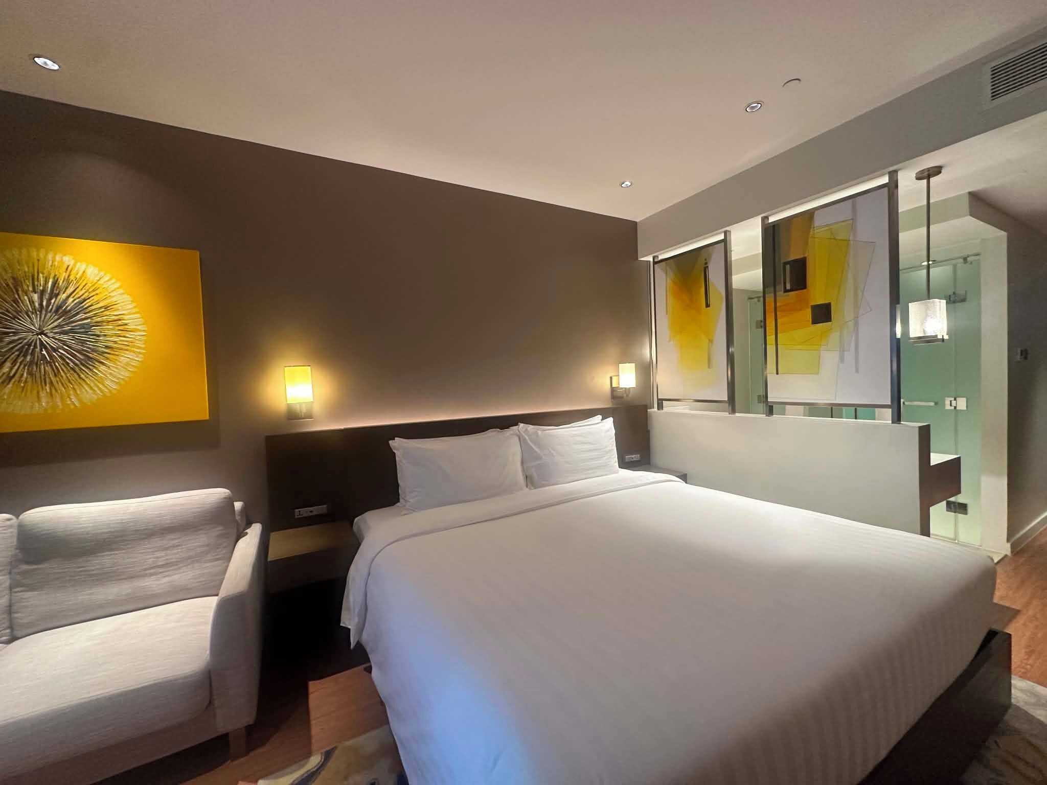 A hotel room with a bed, two bedside tables, a chair, a large mirror, and a wall art piece.