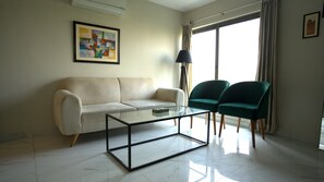 Ein Wohnzimmer mit einer Couch, zwei Sesseln, einem Glastisch, einer Lampe, einem Gemälde und einem großen Fenster mit Vorhängen.