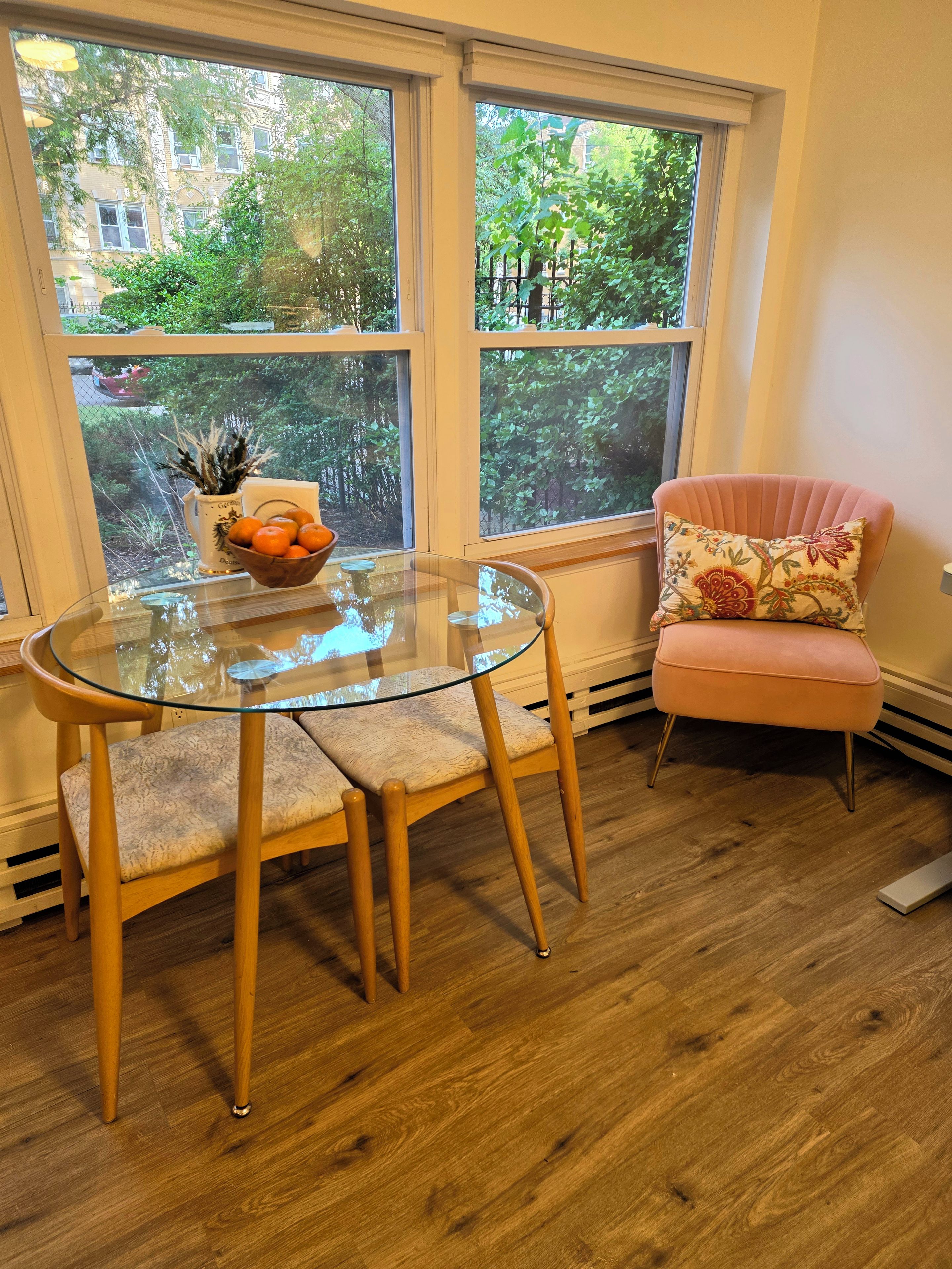 Ein Zimmer mit einem runden Glastisch, einem gepolsterten Sessel und einem Fenster mit Blick auf Grünflächen.