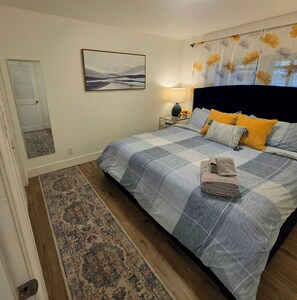 Une chambre à coucher avec un lit, une table de chevet, une lampe, un tableau et une fenêtre avec des rideaux.