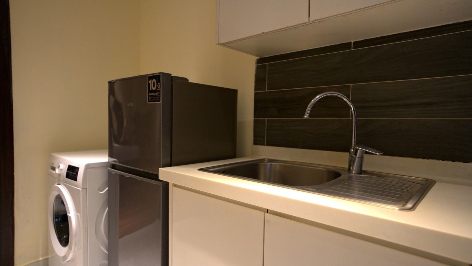 Cocina compacta con refrigerador de acero inoxidable, lavadora integrada y un lavamanos moderno con grifo elegante.