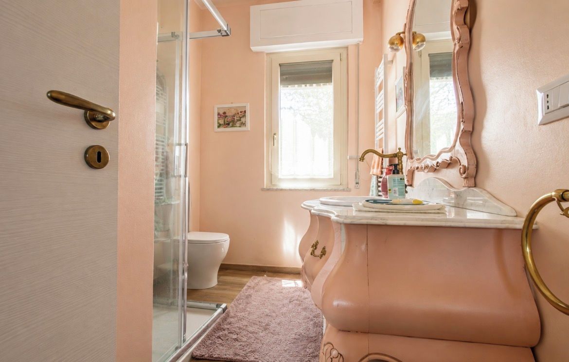 Une salle de bain avec une douche à parois de verre, des toilettes blanches, un meuble-lavabo rose avec un robinet doré et un grand miroir.