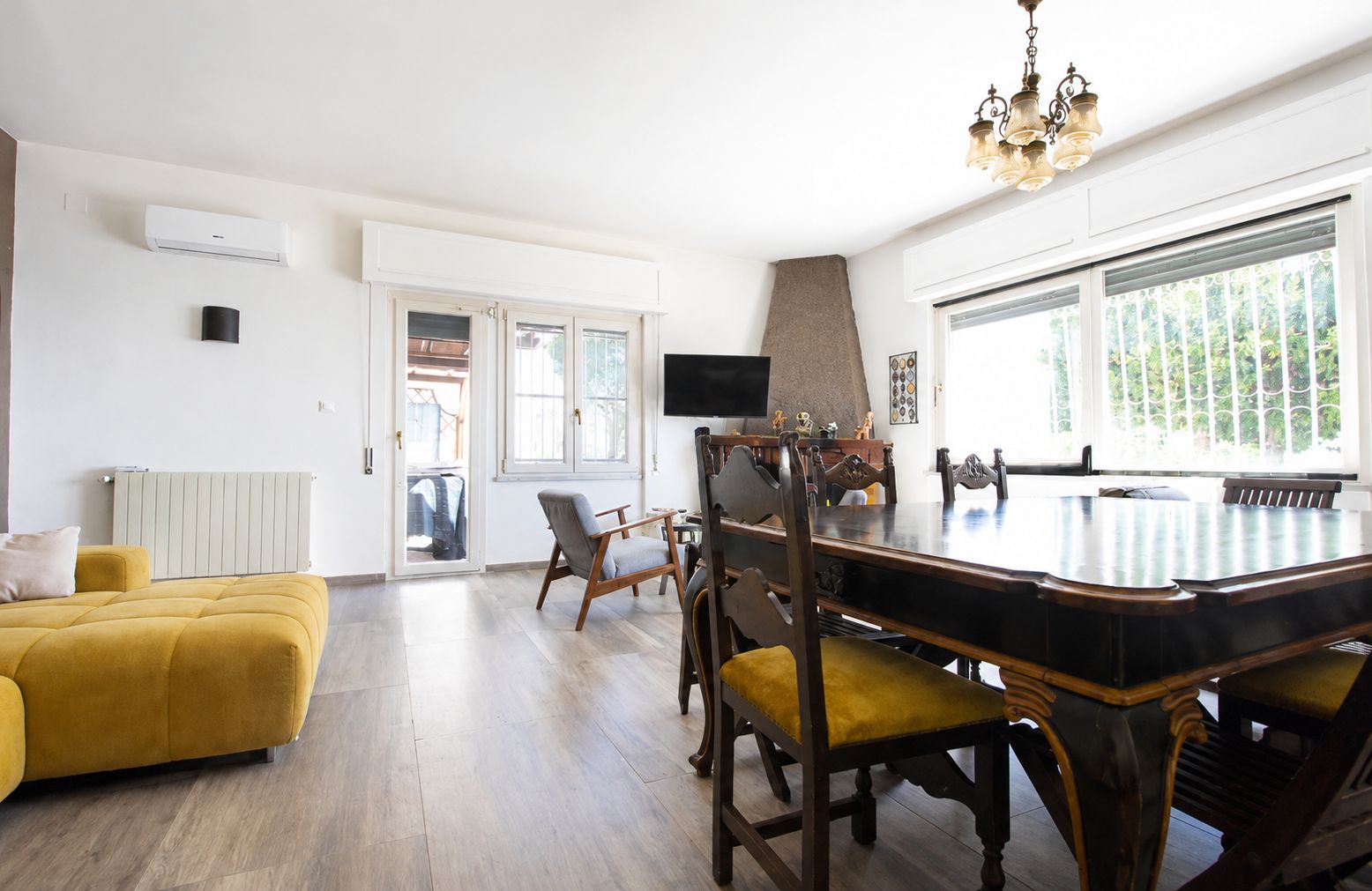 Un vaste espace de vie comprenant un canapé jaune, une table à manger avec des chaises, un téléviseur et une grande fenêtre donnant sur un paysage verdoyant.