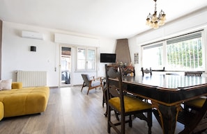 A spacious living area with a yellow sofa, a dining table with chairs, a TV, and a large window with a view of greenery.
