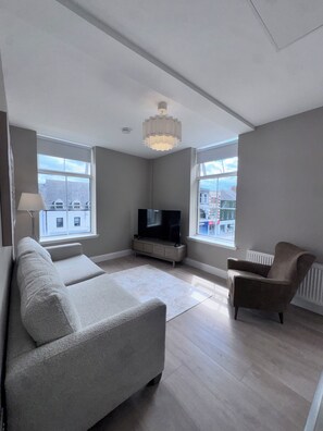 A modern living room with a large grey sofa, a television, and a chair. There are two windows providing a view of the outside.