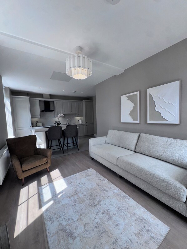 A modern living room with a sofa, a chair, and a rug. There is a kitchen in the background with cabinets and a dining area.