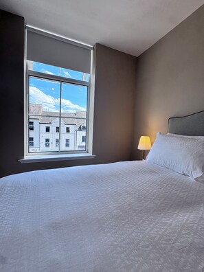 A hotel room with a large window, a bed with white bedding, and a lamp on the nightstand.