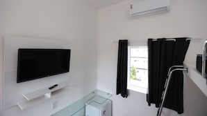 A hotel room with a flat-screen TV, a glass cabinet, a window with curtains, and an air conditioning unit. - A5 suites (Ubatuba)