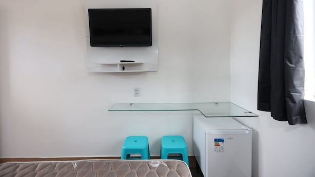 A room with a flat-screen TV mounted on the wall, a glass-top desk, two turquoise stools, and a compact refrigerator. - A5 suites (Ubatuba)