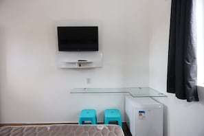 A bedroom with a bed, a TV mounted on the wall, a small fridge, and a glass shelf.