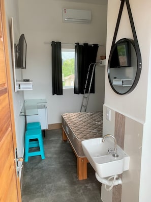Een kleine kamer met een bed, een bureau, een wastafel en een aan de muur bevestigde televisie.