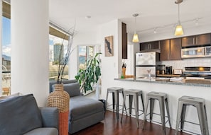 A modern kitchen with dark cabinets, a stainless steel refrigerator, and a microwave. There are three bar stools and a large window offering a view of the city. - Bright 1.5 Bed w/Pool, Parking, HotTub, Sauna, Gym (Vancouver)